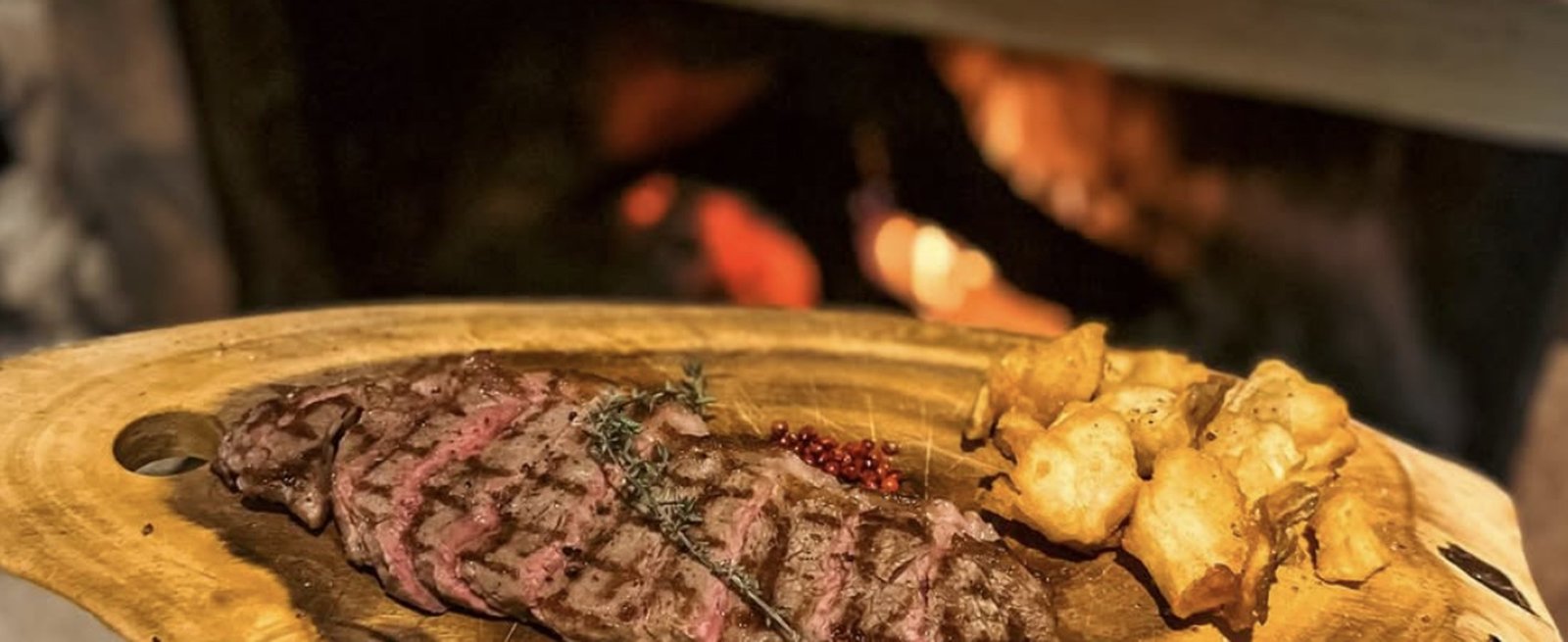 A grilled steak, sliced on a wooden board with roast potatoes, in front of an open fire