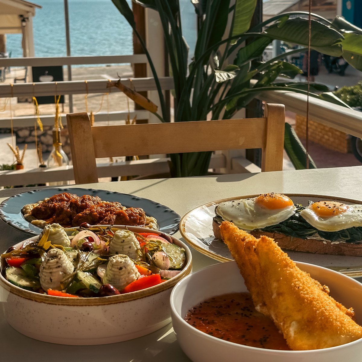 Soutzoukakia, Greek salad, fried Sfela and eggs on sourdough on a table by the window, the sea beyond