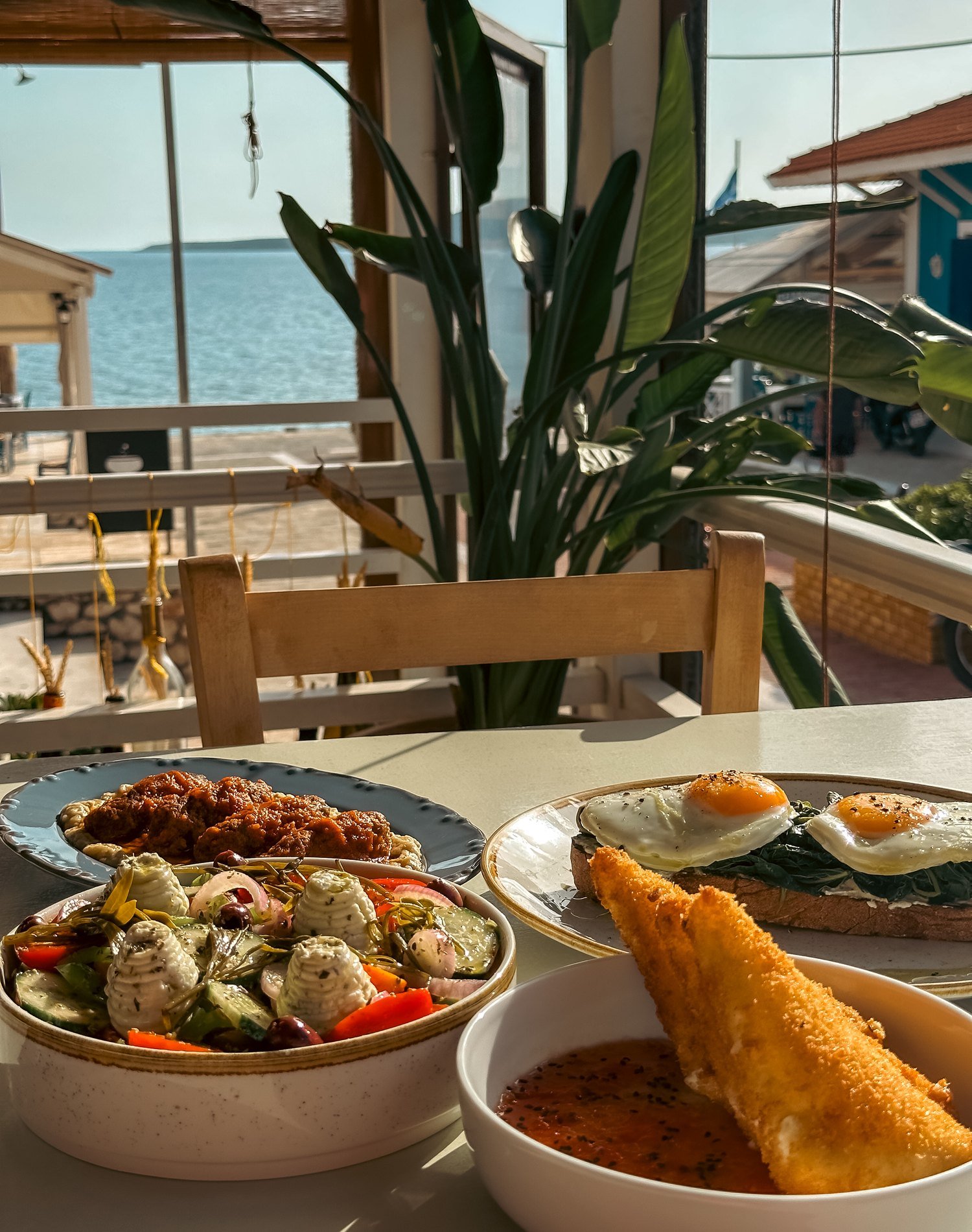 Soutzoukakia, Greek salad, breaded Sfela and eggs on greens, on a table by the window with the sea beyond